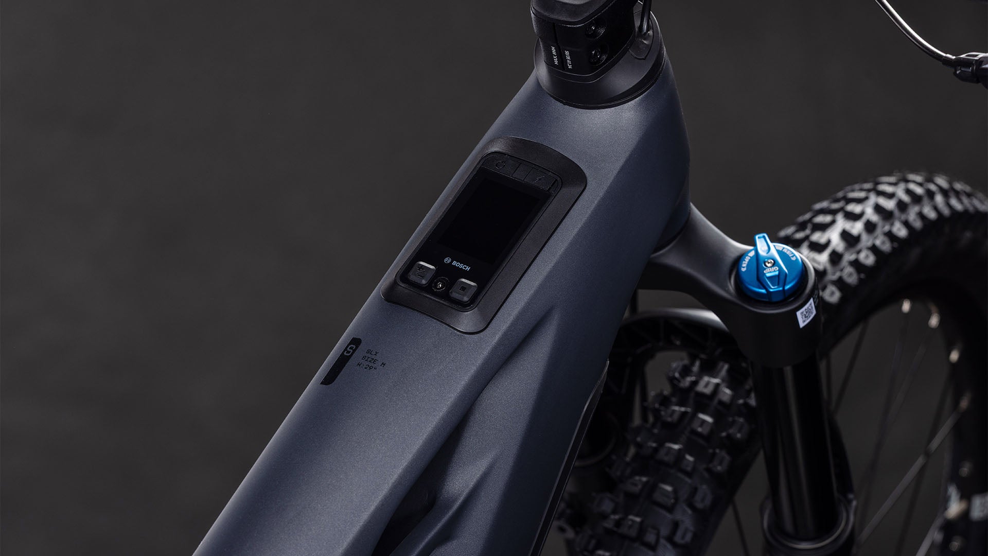 Nahaufnahme des CUBE Stereo Hybrid ONE44 HPC SLX 800 slabgrey'n'orange 2026 E-Mountainbike-Rahmens mit Digitalanzeige, Bedienelementen, Federgabel, Noppenreifen und integriertem Bosch CX-Motor vor dunklem Hintergrund.