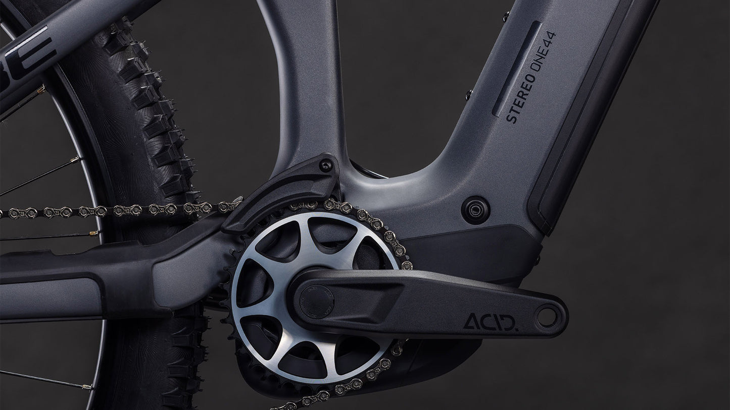 Nahaufnahme der CUBE Stereo Hybrid ONE44 HPC SLX 800 slabgrey'n'orange 2026 Kurbelgarnitur und Kette, mit Shimano XT Schaltung und Bosch CX Motor auf dem schlanken Rahmen mit STEREO ONE44 und ACID Aufschriften vor einem dunklen Hintergrund.