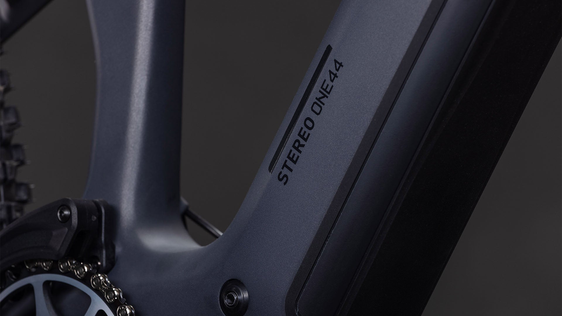 Nahaufnahme des CUBE Stereo Hybrid ONE44 HPC SLX 800 slabgrey'n'orange 2026 Rahmens mit STEREO ONE44 Branding, die einen Teil der Kette und Kurbel zeigt. Mit schlankem Design, glatter Oberfläche und Shimano XT 12-fach Schaltung für präzise Schaltvorgänge.