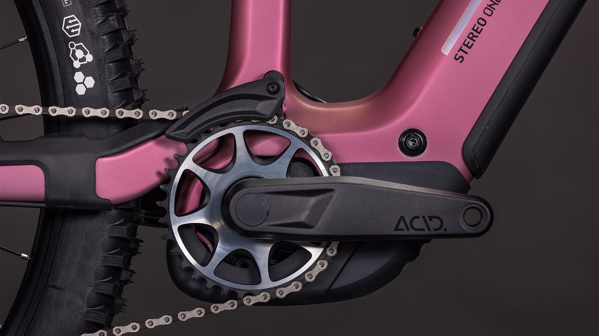 Nahaufnahme der CUBE Stereo Hybrid ONE44 HPC SLX 800 shiftblush'n'art 2026 Kurbelgarnitur, des ACID-gekennzeichneten Pedalarms und des Kettenblatts, wobei ein Teil des Hinterradreifens und des Rahmenlogos zu sehen ist, was die fortschrittliche Integration des Bosch CX-Motors zeigt.
