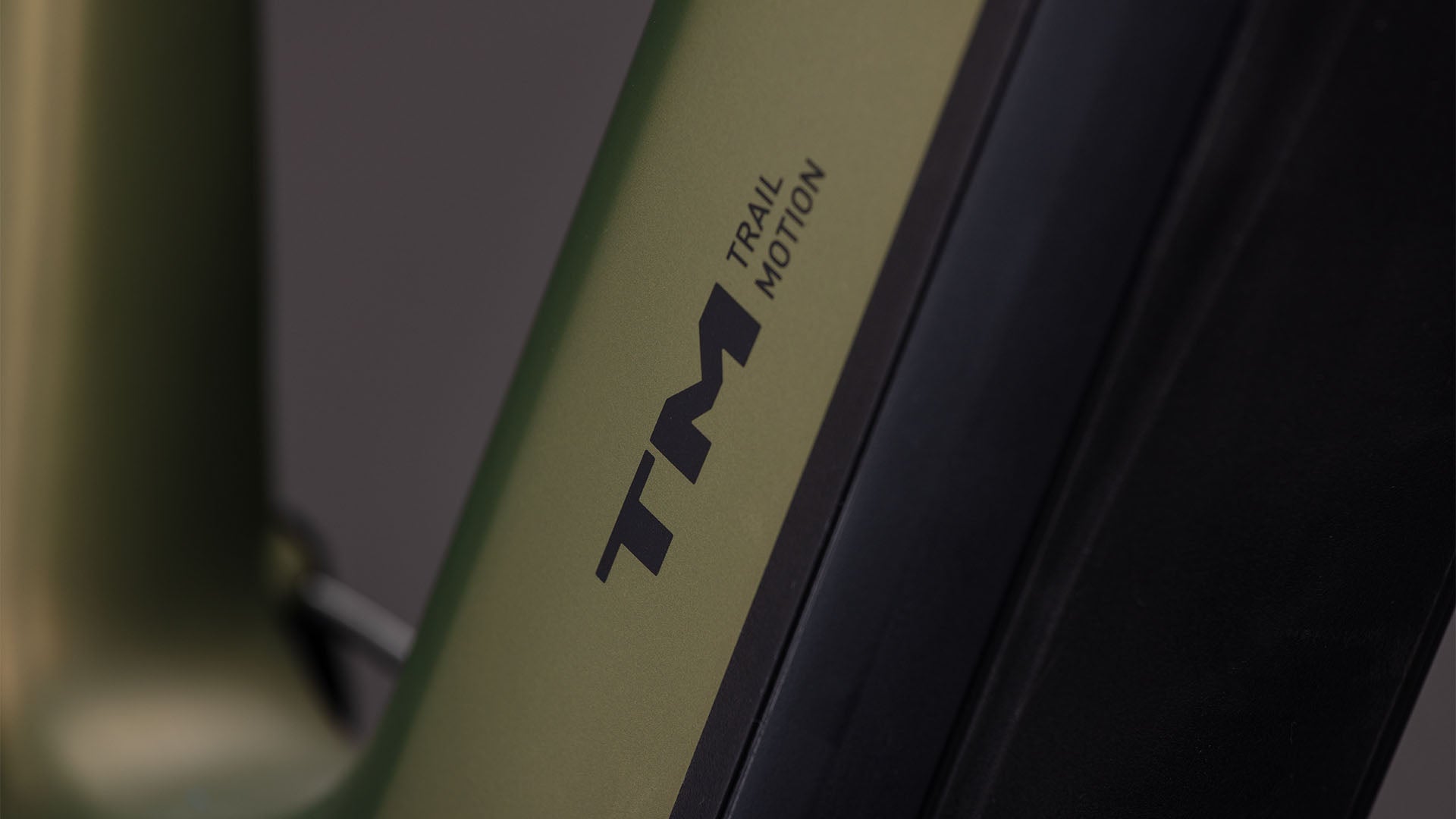 Nahaufnahme eines CUBE Stereo Hybrid ONE44 HPC TM 800 reedgreen'n'matrix 2026 E-Mountainbike mit mattgrünem und schwarzem Rahmen, auf dem der Text TM TRAIL MOTION in schwarzer Schrift zu lesen ist. Der Hintergrund ist unscharf und grau.