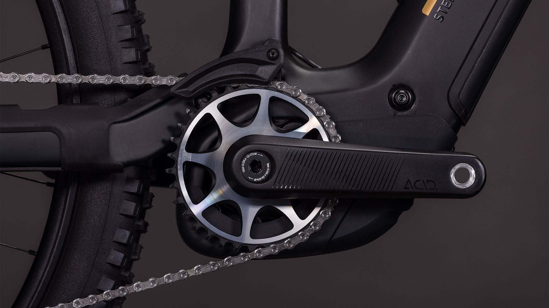 Nahaufnahme des Cube Stereo Hybrid ONE44 HPC AT 800 carbon'n'gold 2026 von CUBE, die die Kurbelgarnitur, das Kettenblatt, die Kette und den Pedalarm in der Nähe des Hinterrads vor einem dunklen Hintergrund zeigt - mit dem fortschrittlichen Bosch CX Motor für nahtlose Leistung.