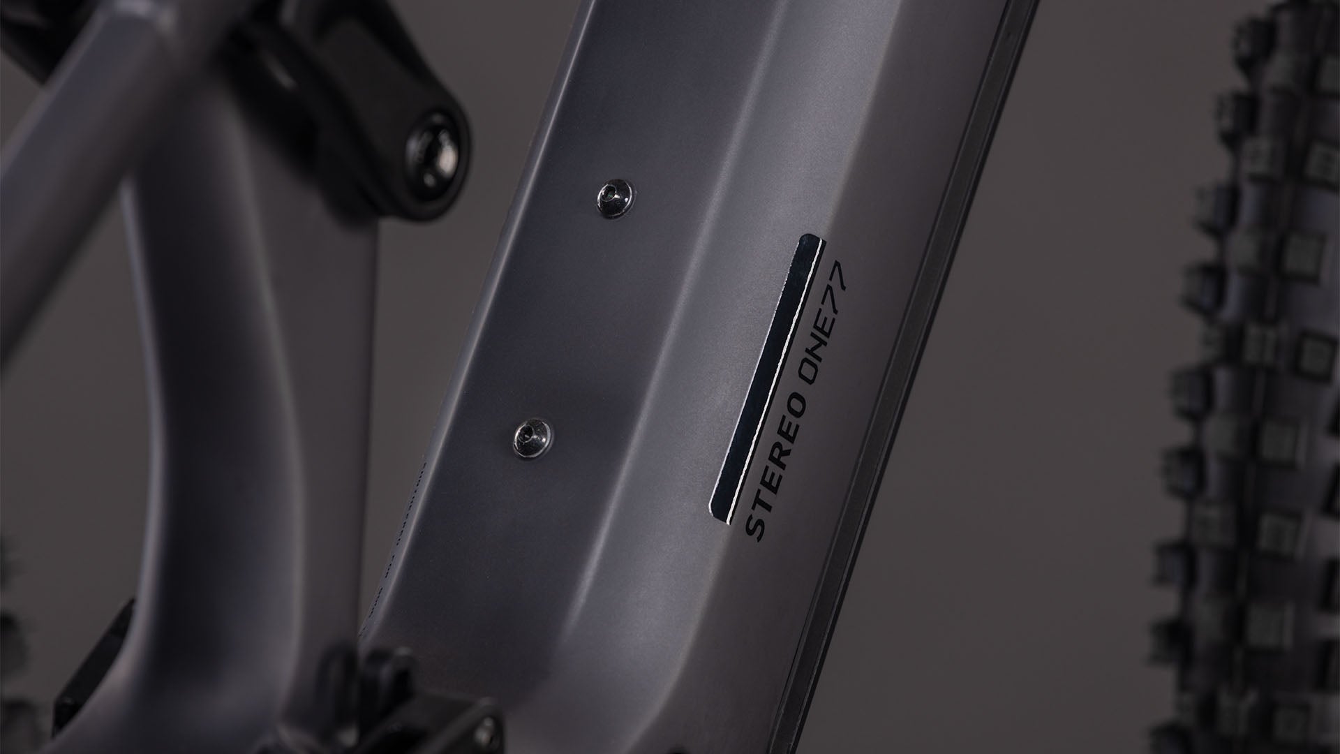 Nahaufnahme des CUBE Stereo Hybrid ONE77 HPC Race 800 in irongrey'n'black, mit dem STEREO ONE77-Logo auf dem Unterrohr, Hinterradfederung, Noppenreifen und Shimano XT 12-fach Schaltung im Hintergrund.