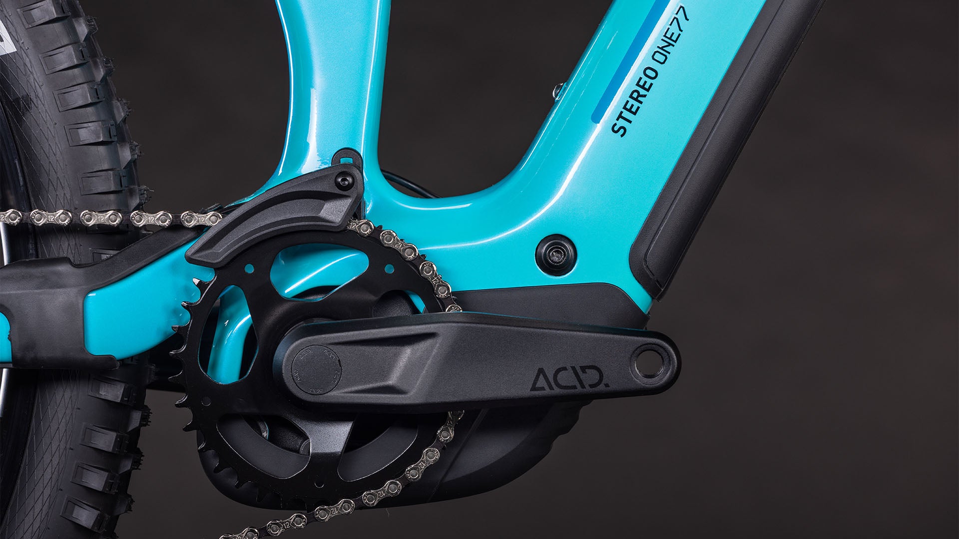 Nahaufnahme des CUBE Stereo Hybrid ONE77 HPC Race 800 swimmingpool'n'black 2026 Rahmens mit der Aufschrift STEREO ONE77. Hervorzuheben sind die Kurbelgarnitur, die Kette und der Pedalarm mit Shimano XT 12-fach Schaltung vor einem schwarzen Hintergrund.
