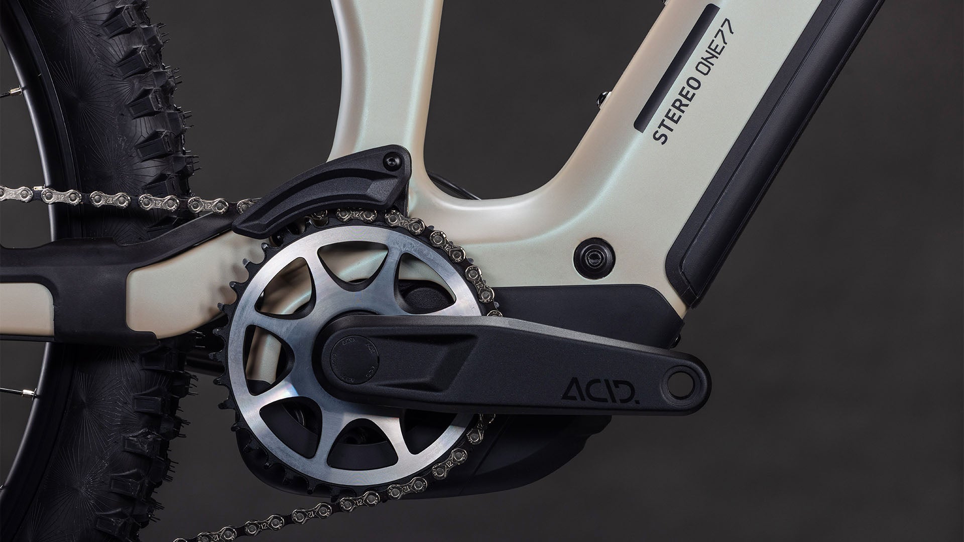 Nahaufnahme einer CUBE Stereo Hybrid ONE77 HPC SLX 800 Kurbelgarnitur, Kettenblatt und ACID Pedalarm am glintsand'n'black Rahmen. Das Bild zeigt auch den knorrigen Hinterreifen, das Kettensegment und die Shimano XT Schaltung.