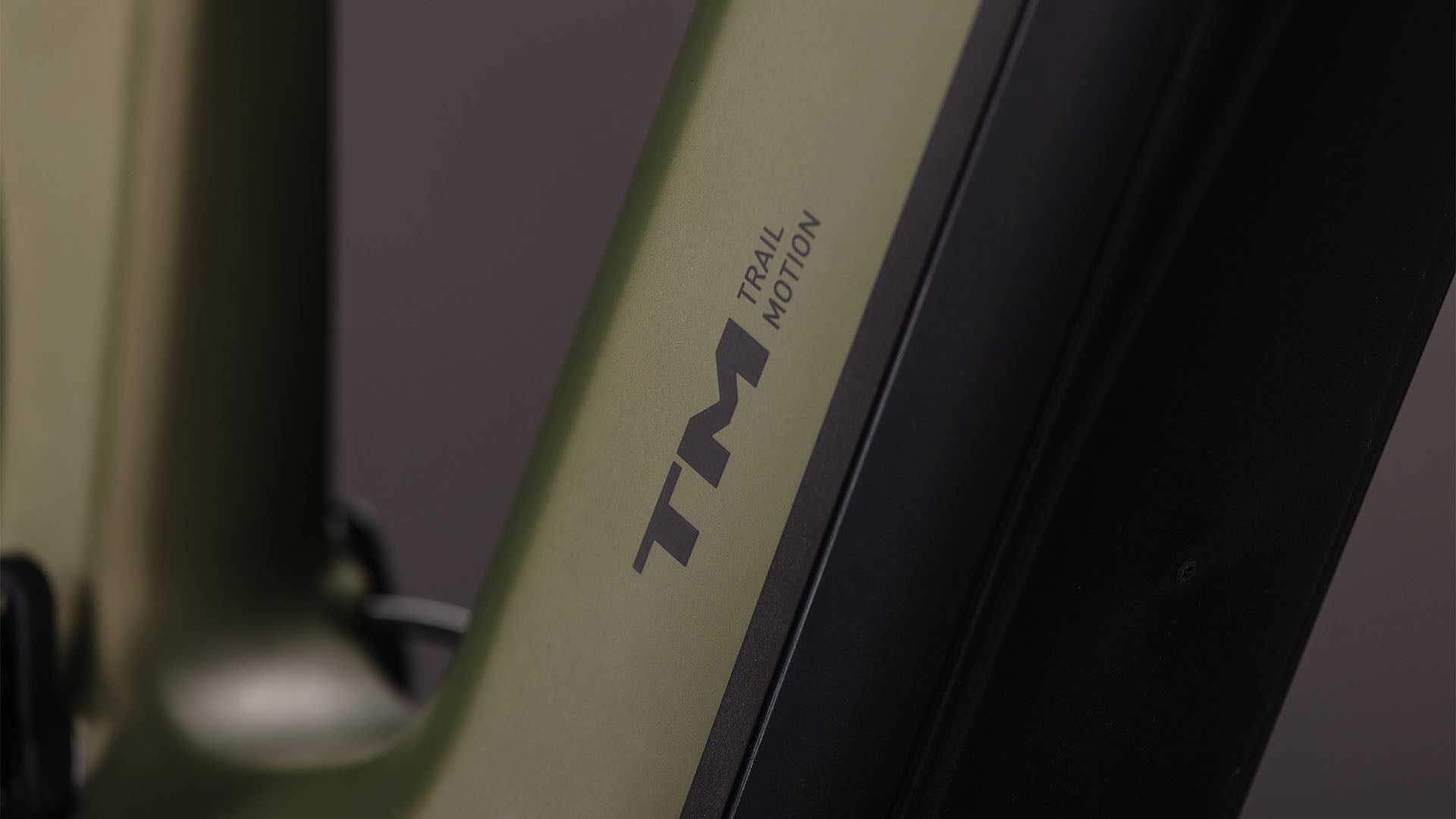 Nahaufnahme des CUBE Stereo Hybrid ONE77 HPC TM 800 reedgreen'n'matrix 2026 Rahmens mit "TM" und "Trail Motion" in schwarz, neben einer schwarzen Sram GX Eagle Getriebekomponente. Der Hintergrund ist unscharf.