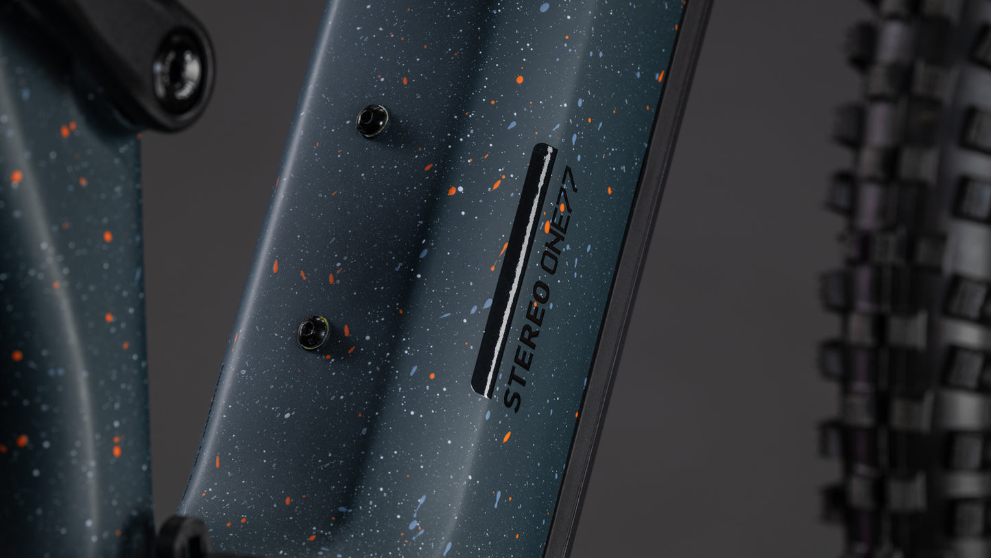 Nahaufnahme des CUBE Stereo Hybrid ONE77 HPC SLT 800 in Galaxy'n'Orange (2026) mit dunkel gesprenkeltem Rahmendesign und dem Schriftzug "STEREO ONE77". Auf der rechten Seite ist ein Stollenreifen zu sehen.