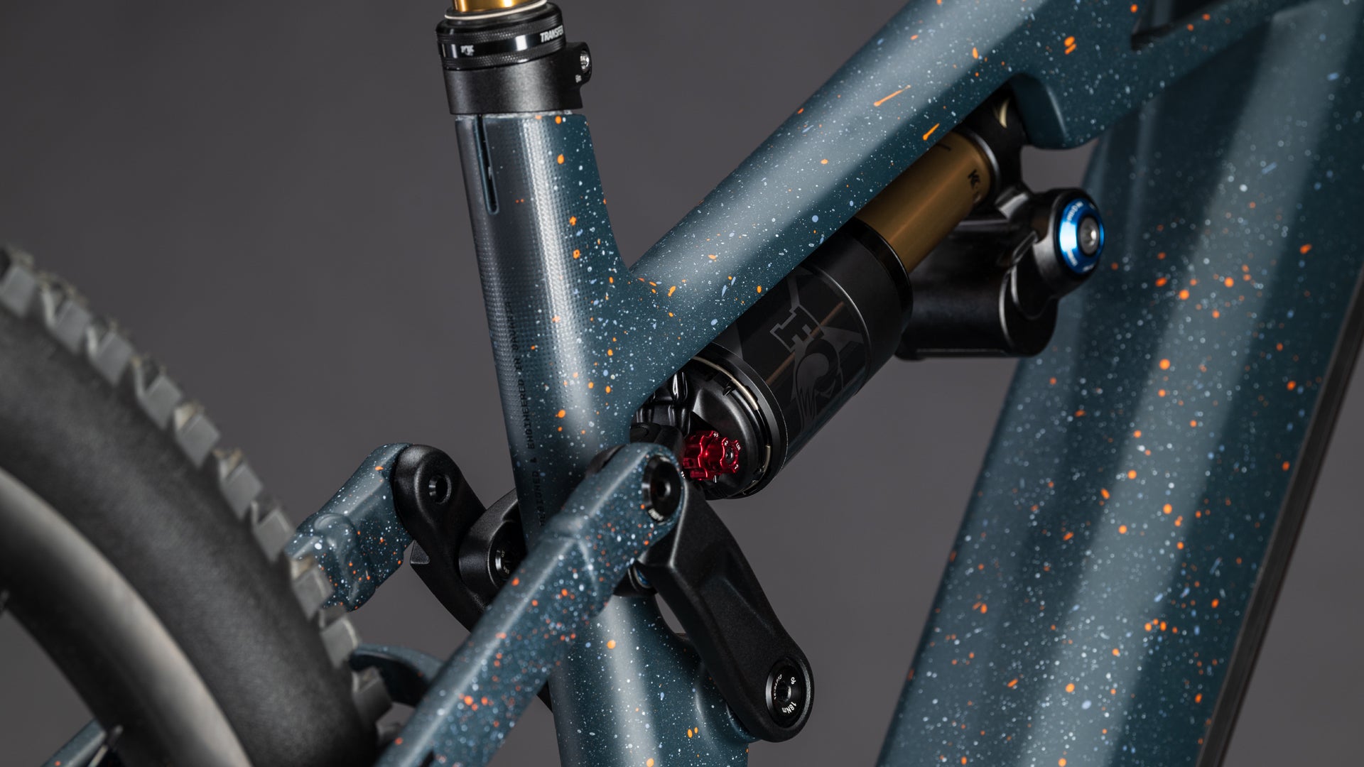 Nahaufnahme der Hinterradaufhängung und des Rahmens des CUBE Stereo Hybrid ONE77 HPC SLT 800 galaxy'n'orange 2026 in mattem Blau mit orangefarbenen Sprenkeln, die den Stoßdämpfer, die Drehpunkte und die Big-Mountain-Performance in der Nähe des Hinterrads hervorheben.