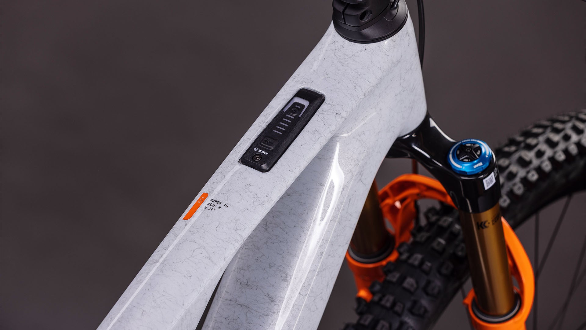 Nahaufnahme eines Cube AMS Hybrid ONE44 C:68X SUPER TM 400X von CUBE in foggrey'n'fibre, die den Carbonrahmen mit elektronischem Bedienfeld, die orangefarbene Federgabel, das matte, strukturierte Finish und die Details des Vorderreifens zeigt.