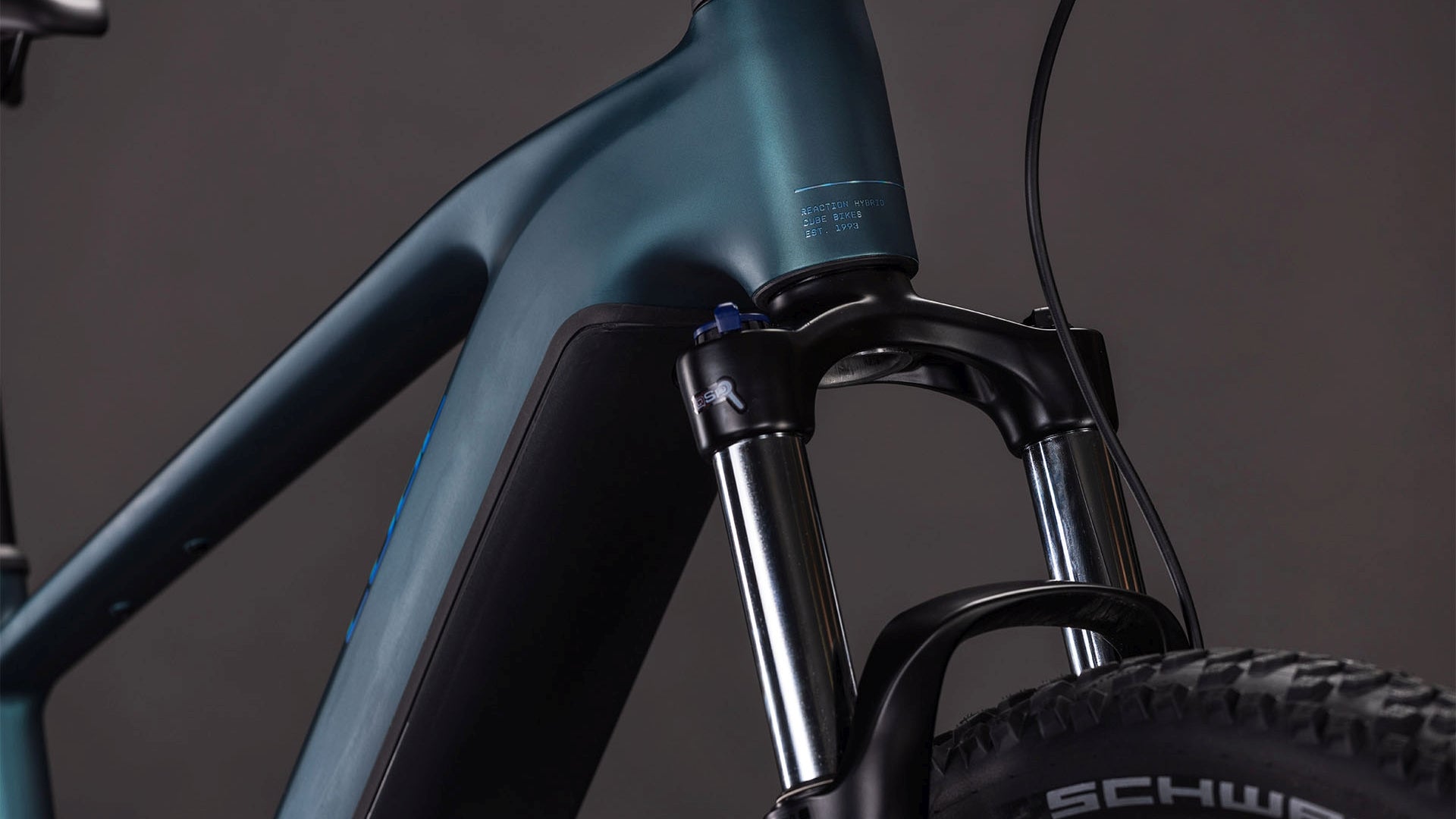 Die Nahaufnahme des CUBE Reaction Hybrid ONE 800 royalgreen'n'prism 2026 zeigt die vordere Federgabel, den blau-grünen Rahmen und die dicken Reifen mit Profil. Der Bosch-Motor hebt die robuste Bauweise und das schlanke Design des Fahrrads vor einem dunklen Hintergrund hervor.