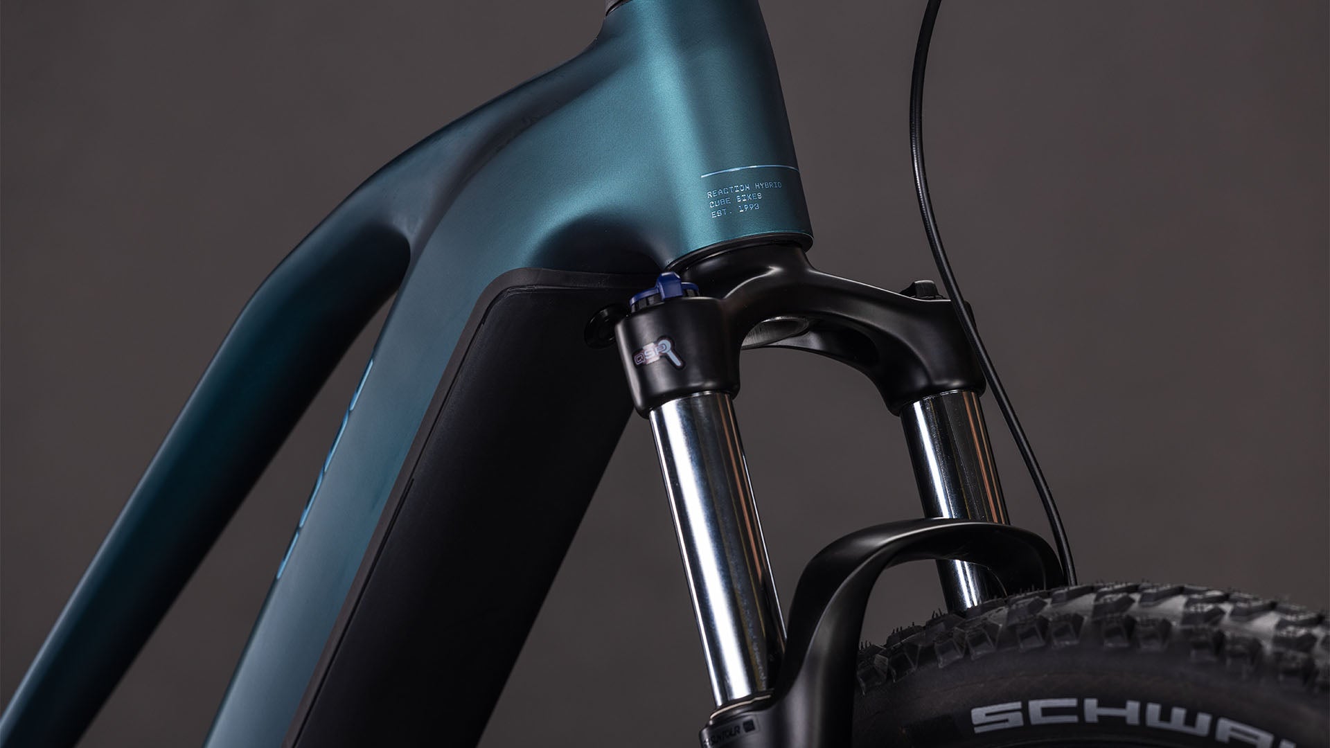 Nahaufnahme des CUBE Reaction Hybrid ONE 800 Trapeze 2026 in royalgreen'n'prism, die die vordere Federgabel, den Bosch-Motor, den Stoßdämpfer und einen Teil des Reifens vor einem dunklen Hintergrund zeigt.