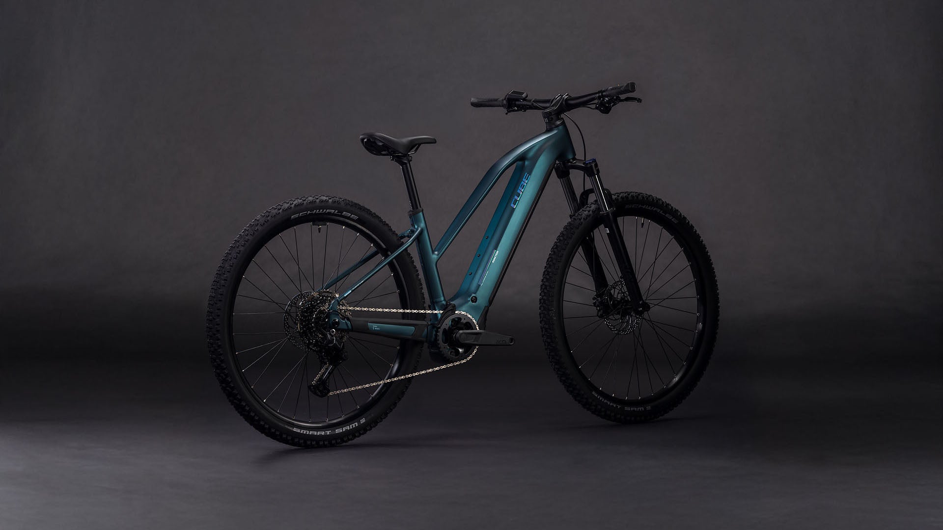 Das CUBE Reaction Hybrid ONE 800 royalgreen'n'prism Trapeze 2026 E-Bike verfügt über einen Bosch-Motor, breite Reifen, Scheibenbremsen, einen stabilen Rahmen und einen schwarzen Sattel, der vor einem dunklen, nahtlosen Hintergrund steht.