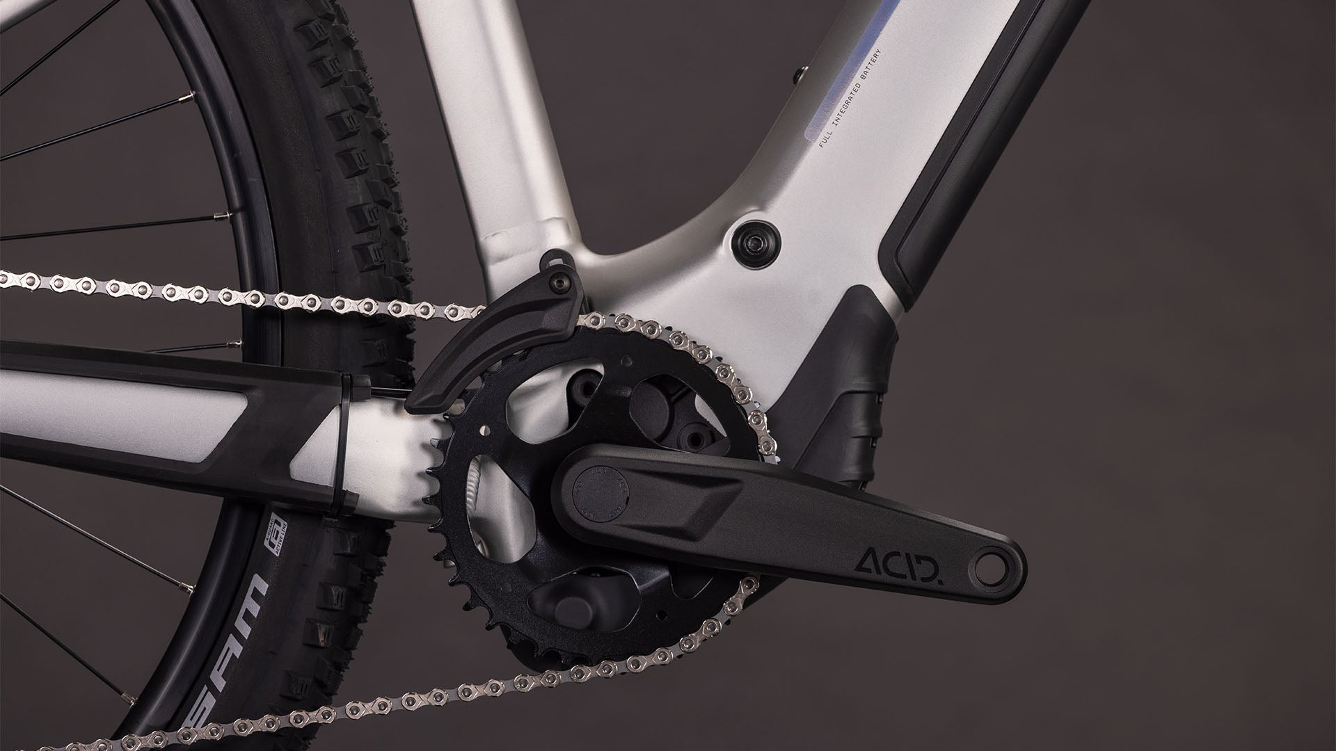 Nahaufnahme eines CUBE Reaction Hybrid ONE 800 sleekgrey'n'prism 2026 Kurbelsatz, Kette und Rahmen mit ACID-gekennzeichnetem Pedalarm und sichtbarem Bosch-Motor. Der dunkle Hintergrund betont die herausragenden Komponenten dieses E-Bikes.