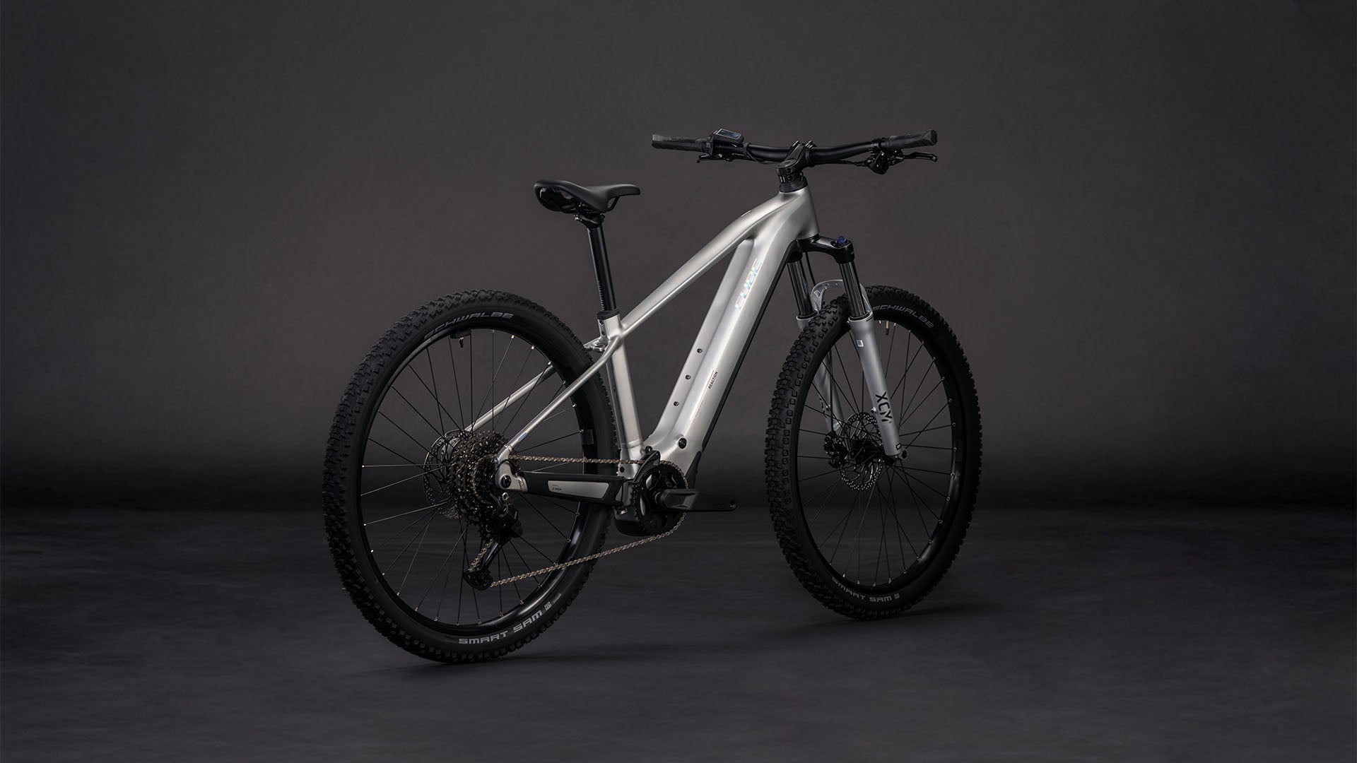 Das CUBE Reaction Hybrid ONE 800 sleekgrey'n'prism 2026 Mountainbike mit dicken Stollenreifen, mattsilbernem Rahmen und Bosch-Motor ist auf einem dunkelgrauen Hintergrund mit Front- und Seitenansicht abgebildet.