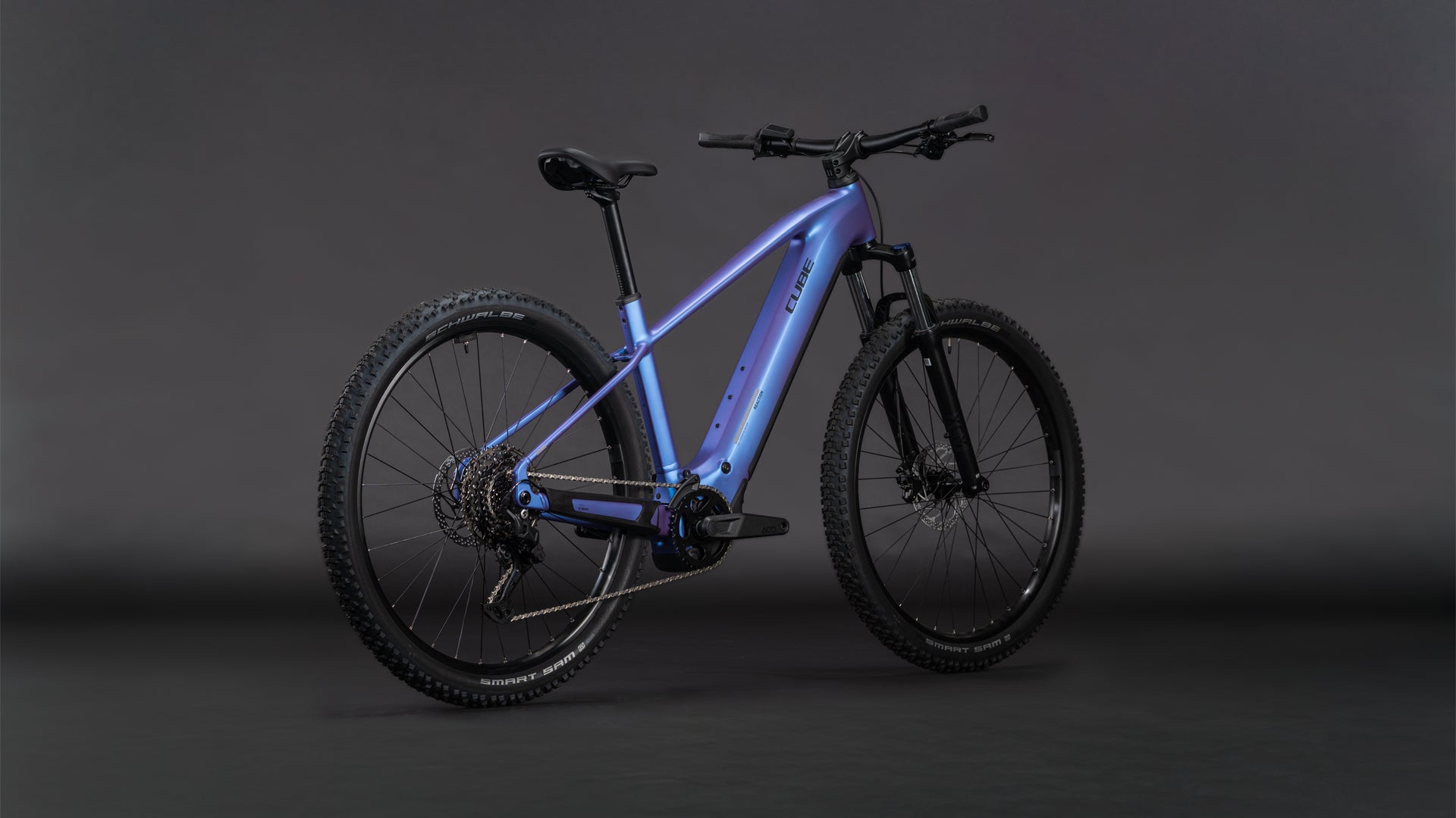 Das CUBE Reaction Hybrid ONE 800 blueiris'n'reflect 2026 Elektro-Mountainbike mit Bosch-Motor, metallisch blauem Rahmen, dicken Stollenreifen und eingebautem 800-Wh-Akku steht vor einem dunklen Hintergrund.
