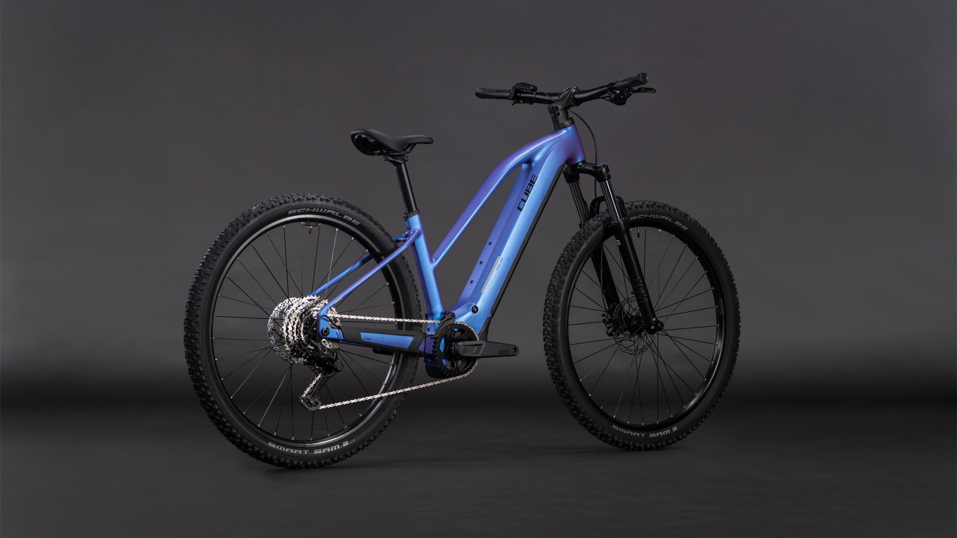 Das CUBE Reaction Hybrid ONE 800 blueiris'n'reflect Trapeze 2026 verfügt über einen metallisch blauen Rahmen, dicke Offroad-Reifen, einen Bosch-Motor, einen 800-Wh-Akku, eine Federgabel, Scheibenbremsen und einen aufrechten Sitz auf einem dunklen, neutralen Hintergrund.