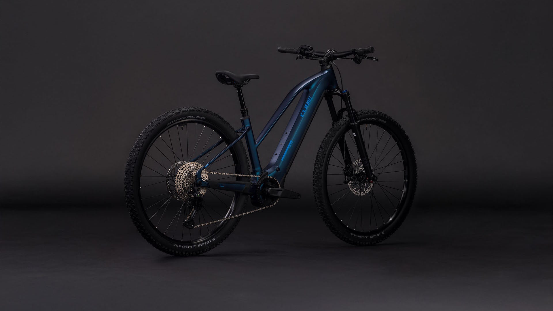 Das CUBE Reaction Hybrid Pro 800 nebular'n'dazzle Trapeze 2026 ist ein blaues E-Mountainbike mit dicken Reifen, Scheibenbremsen, Durchstiegsrahmen und einem 800-Wh-Akku am Unterrohr, der vor einem dunklen Hintergrund steht.
