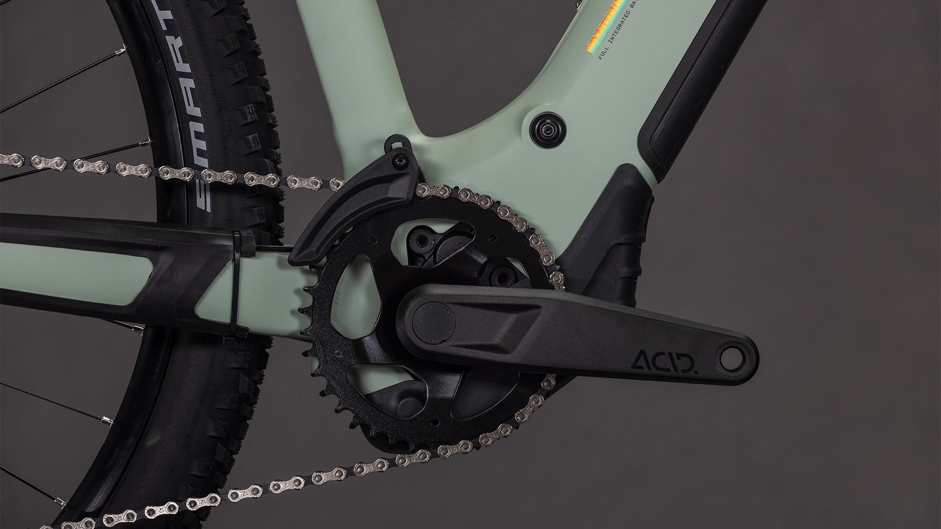 Nahaufnahme des Cube Reaction Hybrid Pro 800 desertstone'n'driedherbs Trapeze 2026 von CUBE: Kurbelsatz und Kette auf einem hellgrünen E-MTB-Rahmen mit Bosch CX Antrieb, ACID-gekennzeichnetem Kurbelarm und Hinterradreifen im Hintergrund.