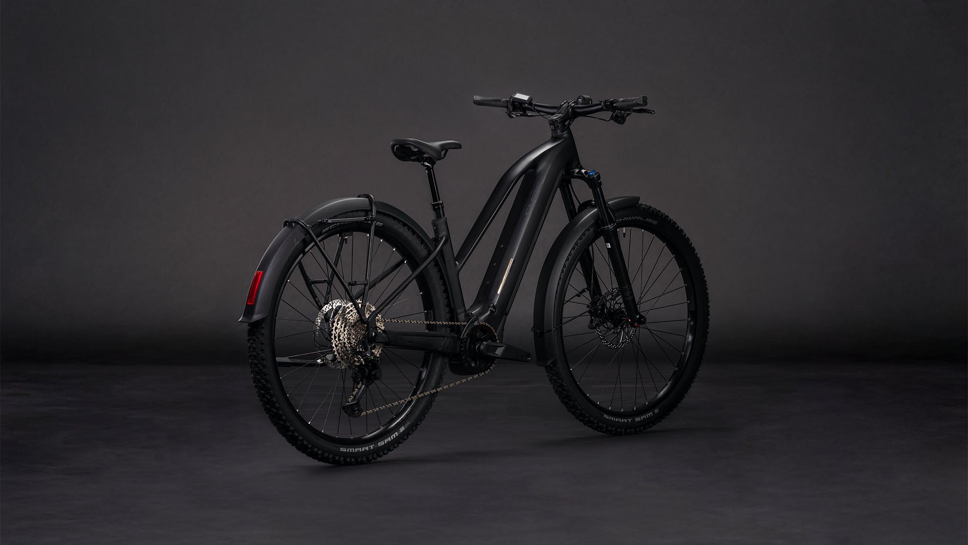 Das CUBE Reaction Hybrid Pro 800 FE blackline Trapeze 2026, ein mattschwarzes E-Bike mit dicken Reifen, Hinterradschutzblech und Scheibenbremsen, ausgestattet mit einem Bosch CX Motor, wird in einem Studio mit dunklem Hintergrund ausgestellt und von der Seite und von hinten gezeigt.