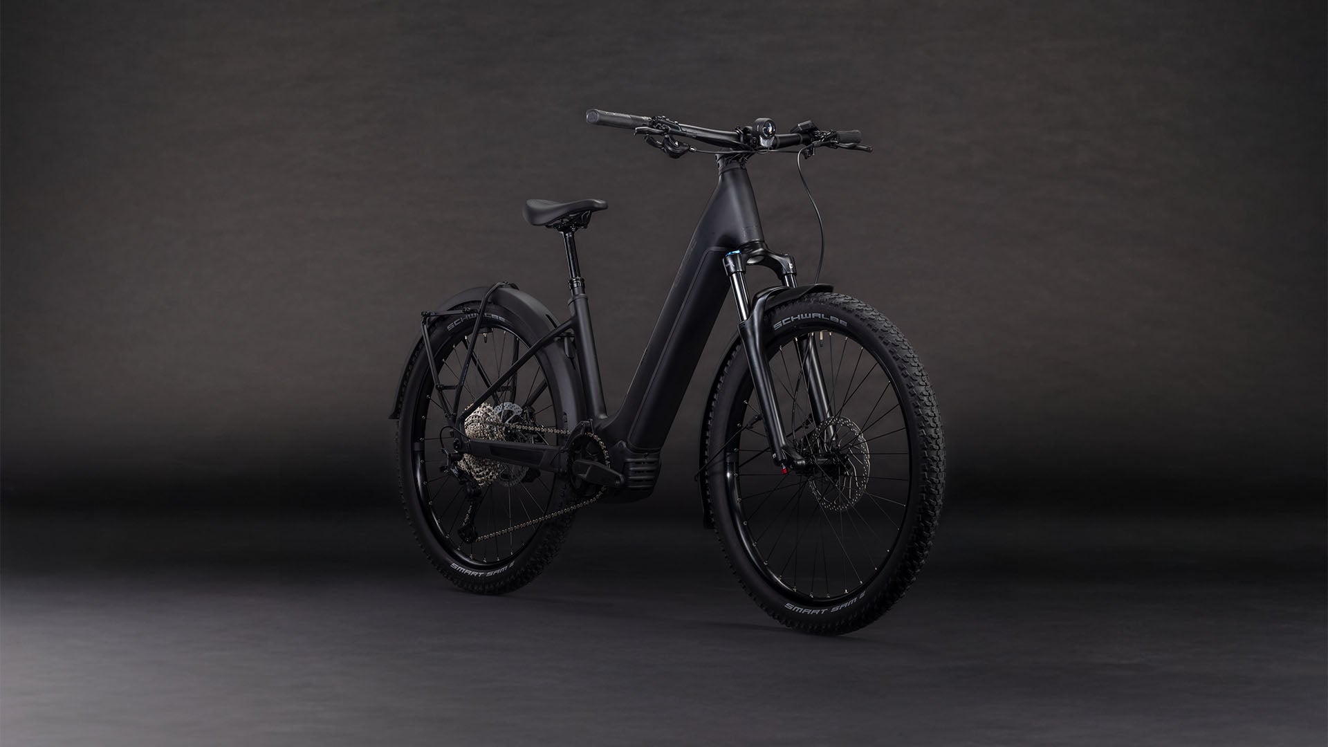 Das CUBE Reaction Hybrid Pro 800 FE blackline Easy Entry 2026, ein mattschwarzes E-Bike mit dicker Bereifung, Federgabel, Schutzblechen und Durchstiegsrahmen, besticht durch einen Bosch CX-Motor und modernes Design vor dunklem Hintergrund.