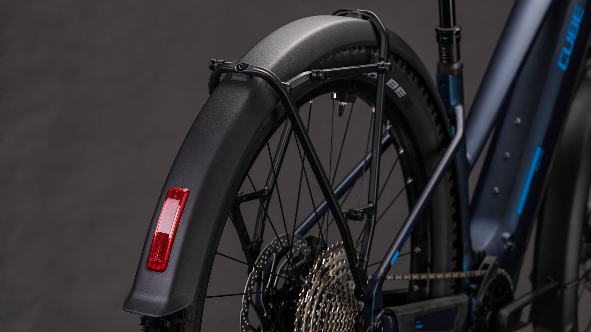 Nahaufnahme des Cube Reaction Hybrid Pro 800 FE nebular'n'dazzle Trapeze 2026 von CUBE, die das Hinterrad, das Schutzblech, den Bosch CX-Motor, den Antriebsstrang, die Shimano XT-Schaltung, den Reflektor und den Rahmen vor einem dunklen Hintergrund zeigt.