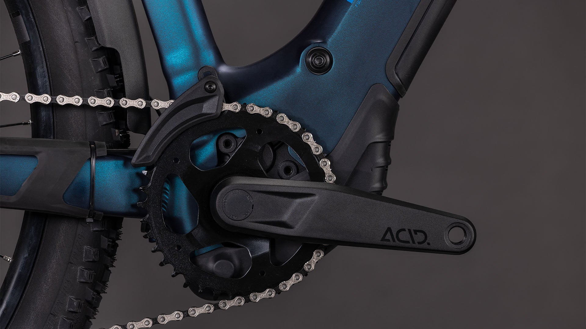 Die Nahaufnahme des CUBE Reaction Hybrid Pro 800 FE nebular'n'dazzle Trapeze 2026 Antriebs zeigt den schwarzen ACID Kurbelarm, das Kettenblatt und die Kette auf einem dunkelblauen Rahmen mit Shimano XT Schaltung vor einem schwarzen Hintergrund.