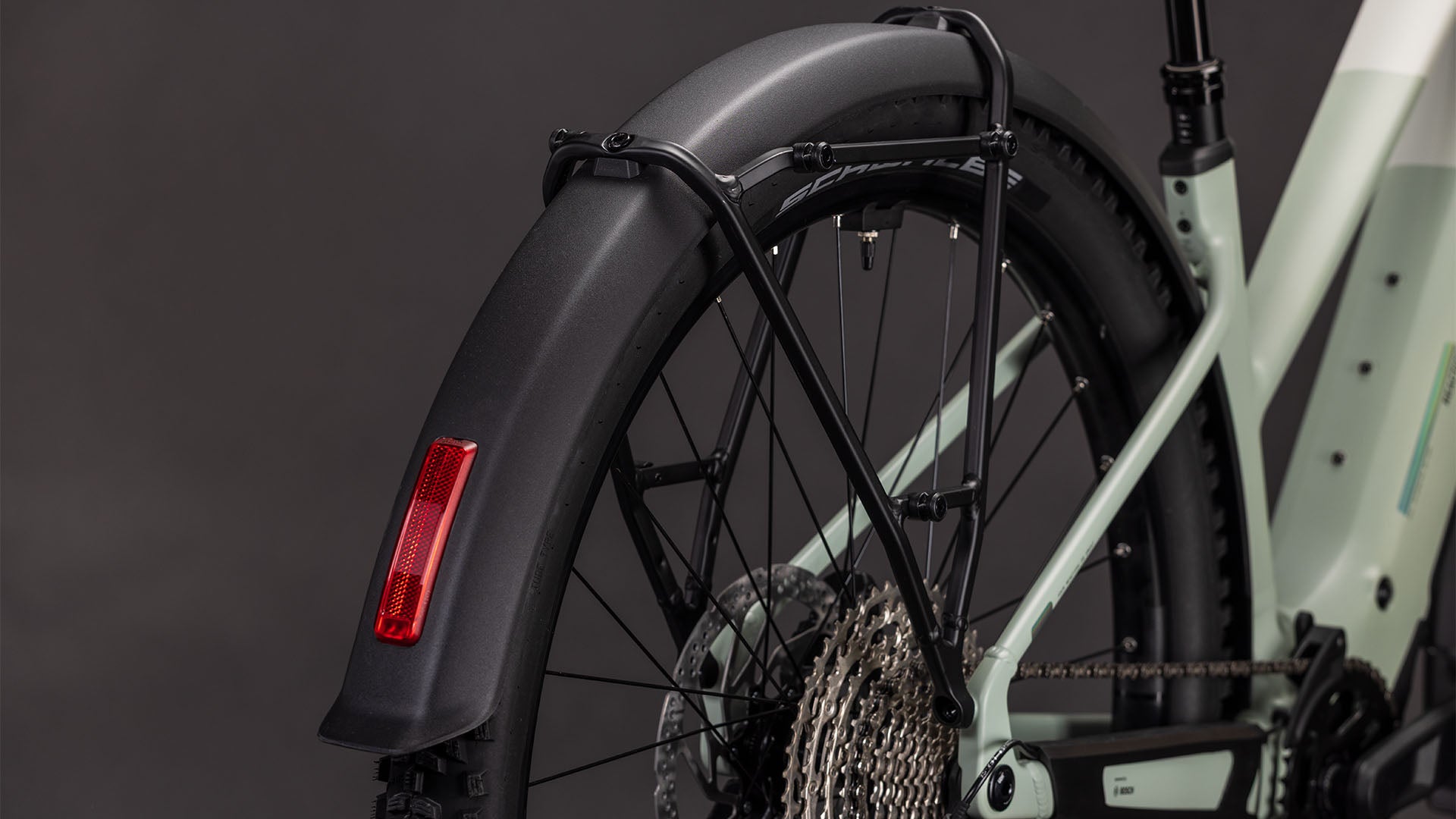 Nahaufnahme des Hinterrads des CUBE Cube Reaction Hybrid Pro 800 FE desertstone'n'driedherbs Trapeze 2026, die das schwarze Schutzblech mit rotem Reflektor, die Shimano XT 12-fach Schaltung und den E-Bike-Rahmen vor einem dunklen Hintergrund zeigt.