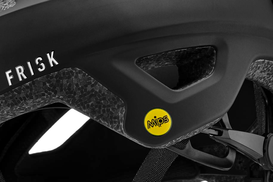 Nahaufnahme des schwarzen CUBE Helm FRISK MTB Trail Helm von Cube mit strukturiertem Schaumstoff, "FRISK"-Schriftzug, gelbem MIPS-Technologie-Aufkleber an der Seite und einem teilweise sichtbaren Riemen mit Schnalle.