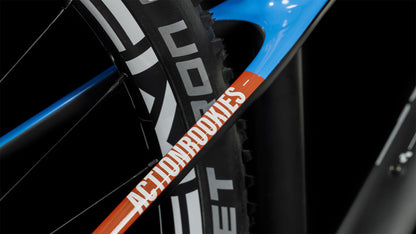 Nahaufnahme des CUBE Elite 240 C:62 Pro carbon'n'blue'n'red 2024 Rahmens mit dem Schriftzug "ACTION ROOKIES" auf einem orangefarbenen Streifen, mit einem schwarzen Reifen und Teilen des blauen und schwarzen CUBE Rahmens im Hintergrund.