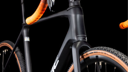 Nahaufnahme des CUBE Cross Race C:62 Pro carbon'n'orange 2025, die den Carbonrahmen, das orangefarbene Lenkerband, die hellbraunen Noppenreifen und die Shimano 105 Di2 Teile hervorhebt. Die Vorderseite des Fahrrads hebt sich von einem schwarzen Hintergrund mit feinen Texturdetails ab.