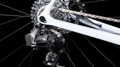 Nahaufnahme des Schaltwerks, der Kette und der Kassette eines CUBE Cross Race C:62 Race polarwhite'n'blue 2025 vor schwarzem Hintergrund. Zu sehen sind Metallteile, Sram Rival AXS-Details und ein Teil des weißen Rahmens.