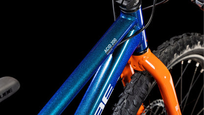Nahaufnahme des CUBE Acid 200 Disc Actionteam 2025 Mountainbikes von CUBE mit metallic-blauem Rahmen, Shimano 7-Gang-Schaltung, orangefarbener Vordergabel und dicken schwarzen Reifen vor schwarzem Hintergrund.