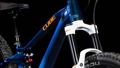 Nahaufnahme eines blauen CUBE Stereo ONE22 Rookie actionteam Rahmens mit orangem Branding, Federgabel, dickem Reifenprofil, Shimano 11-fach Schaltung und einem Teil des Lenkers vor schwarzem Hintergrund.