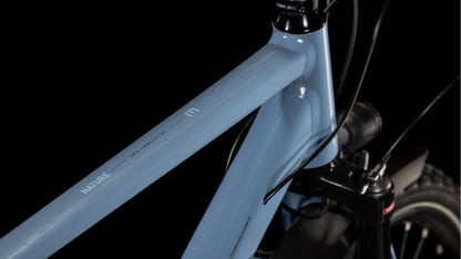 Nahaufnahme eines taubenblauen und schwarzen CUBE Nature EXC Allroad-Rahmens mit "NATURE"-Schriftzug, der eine robuste Konstruktion, klare Linien und eine Shimano Cues 2x11-Gang-Schaltung vor einem schwarzen Hintergrund zeigt.