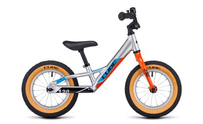 Das Cube Cubie 120 walk actionteam Laufrad ist ein leichtes Aluminium-Balancebike in Silber mit orangefarbener Gabel, schwarzem Sitz, blauem CUBE Branding und beigen Reifen. Es hat keine Pedale oder Stützräder und ist auf einem schlichten weißen Hintergrund abgebildet.