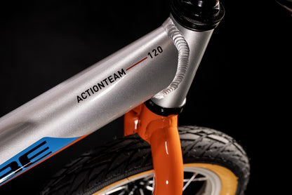 Nahaufnahme des Cube Cubie 120 walk actionteam Laufrad, mit dem ACTIONTEAM 120 auf dem silbermetallicfarbenen Rahmen mit orangefarbener Gabel und schwarzem Lenker - ein leichtes Aluminium-Balancebike für Kinder, vor einem dunklen Hintergrund.
