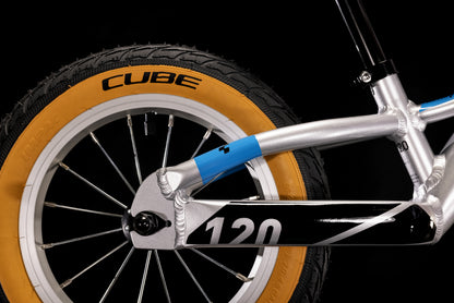 Nahaufnahme des Hinterrads eines Cube Cubie 120 walk actionteam Laufrads mit einem hellbraunen Reifen mit der Aufschrift "CUBE", silbernen Speichen und einem silber-schwarzen Rahmen mit blauen Akzenten vor einem schwarzen Hintergrund.
